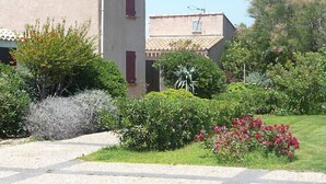 Il s’agit d’un bâtiment à la façade beige, doté d’une porte rouge et d’une petite fenêtre. Un jardin avec diverses plantes et fleurs se trouve devant le bâtiment.