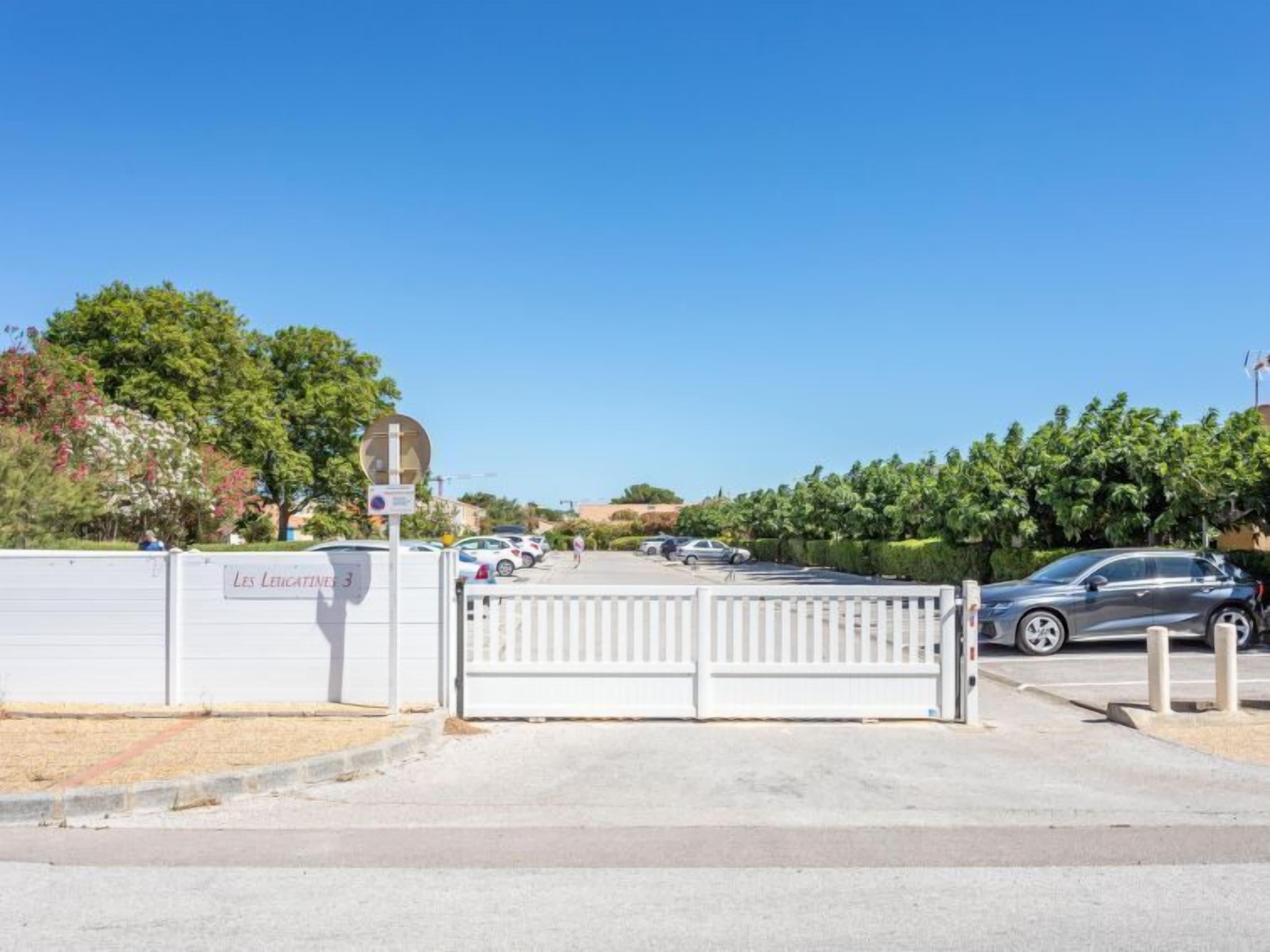 A gated parking area with a white fence, a sign reading "Les Leucatiens 3", and a clear blue sky.