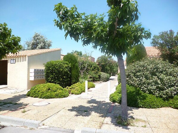 Zona residencial con un camino empedrado, un pequeño edificio con techo de tejas y diversos arbustos y árboles verdes.