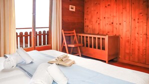 A neatly made bed with towels and an open book, a wooden chair, and a wooden crib in a room with wooden walls and a balcony with a wooden fence. - Vacation home Mungg by Interhome (Nendaz)