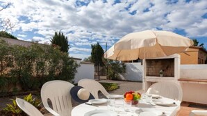 Une terrasse avec une table ronde prévue pour quatre personnes, entourée de chaises en plastique blanches, sous un parasol beige.