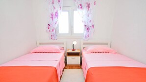 Et soveværelse med to senge, begge med pink sengetøj og orange dyner, et natbord med en lampe, og et vindue med blomstrede gardiner.