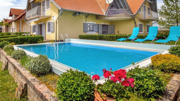 Ein Poolbereich mit einem rechteckigen Pool, Liegestühlen und einem Gebäude mit rotem Ziegeldach und Balkonen.