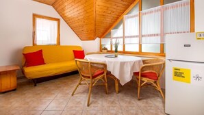 Ein gemütliches Zimmer mit Holzbalkendecke, einem gelben Sofa mit roten Kissen, einem runden Tisch mit weißem Tischlauf, Korbstühle und einem Kühlschrank mit Aufkleber.
