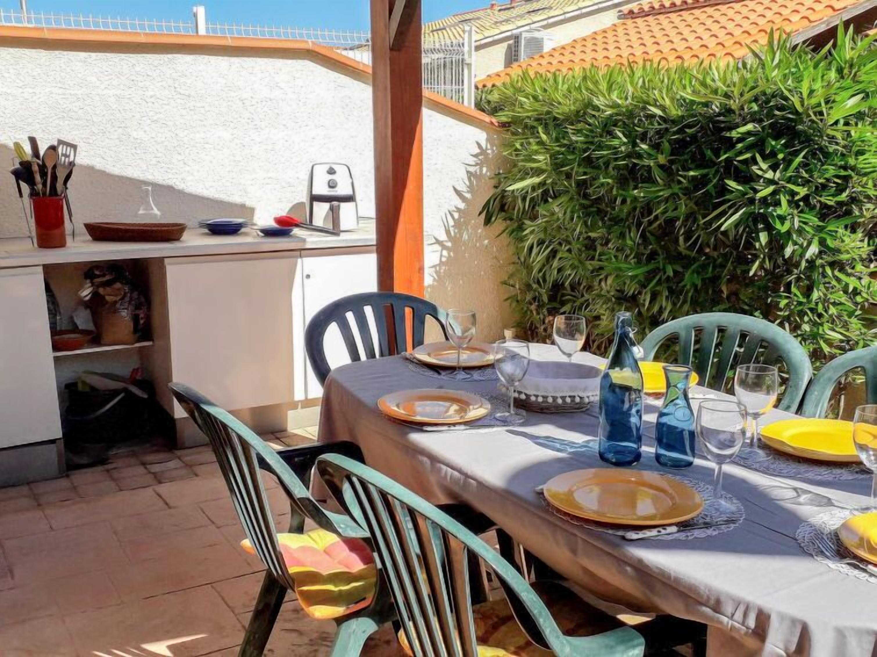 Eine Terrasse mit Esstisch, umgeben von Stühlen, sowie eine kleine Küchenzeile mit Geschirr und Wasserspender.