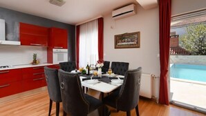 A modern kitchen with red cabinets, a dining area with a white table and black chairs, and a view of a swimming pool through large windows. - Vacation home Jure by Interhome (Zadar)