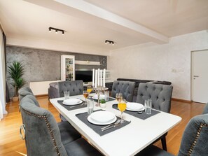 Ein Essbereich mit einem weißen Tisch, gedeckt für vier Personen, grauen Polsterstühlen, einer weißen Kommode mit Flaschen und einem grauen Sofa im Hintergrund.