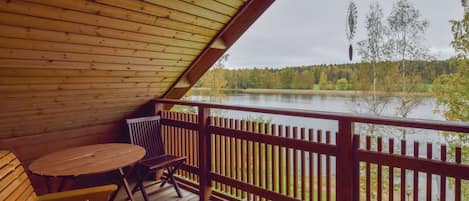 A wooden deck with a table and chairs, overlooking a lake with trees in the background.