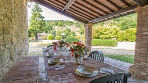 Un espace de restauration extérieur abrité, doté d’un mur en pierre, d’une table en bois dressée avec des assiettes, des verres et des fleurs, et offrant une vue sur un jardin et des collines en arrière-plan.