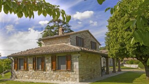 Une maison en pierre avec un toit en tuiles, entourée d’une pelouse bien entretenue et d’arbres.
