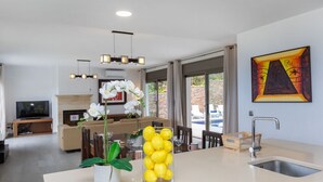 Et moderne køkken med en central ø, et spiseområde med stole, en sofa og et fjernsyn i baggrunden.
