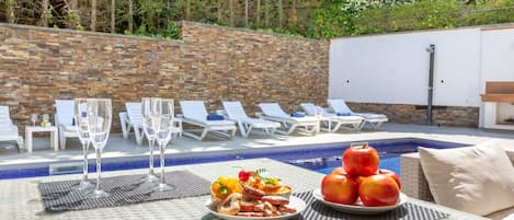 A poolside table with food, glasses, and a fruit display.