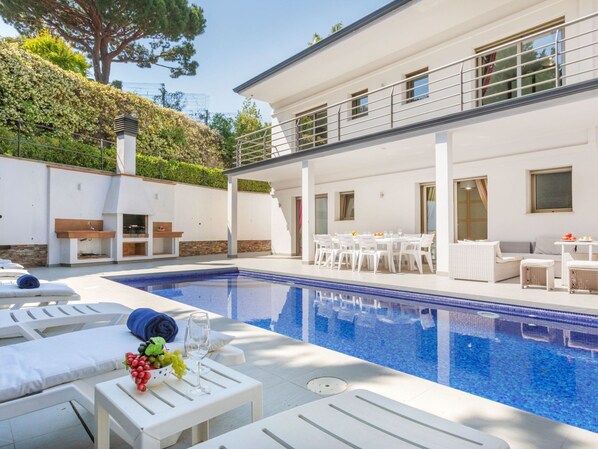 A modern outdoor pool area with lounge chairs, a dining setup, and a white building with a balcony. - Palmbeach by Interhome (Lloret de Mar)