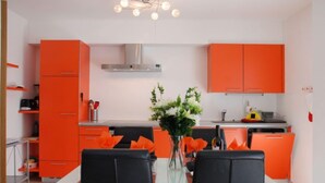 Eine moderne Küche mit orangefarbenen Schränken, einer Edelstahl-Dunstabzugshaube und einem Essbereich mit einem Glastisch und schwarzen Stühlen.