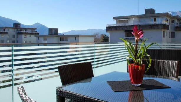 Ein Balkon mit Glastisch und Stühlen, einer roten Vase mit Blumen und Blick auf Gebäude und Berge.