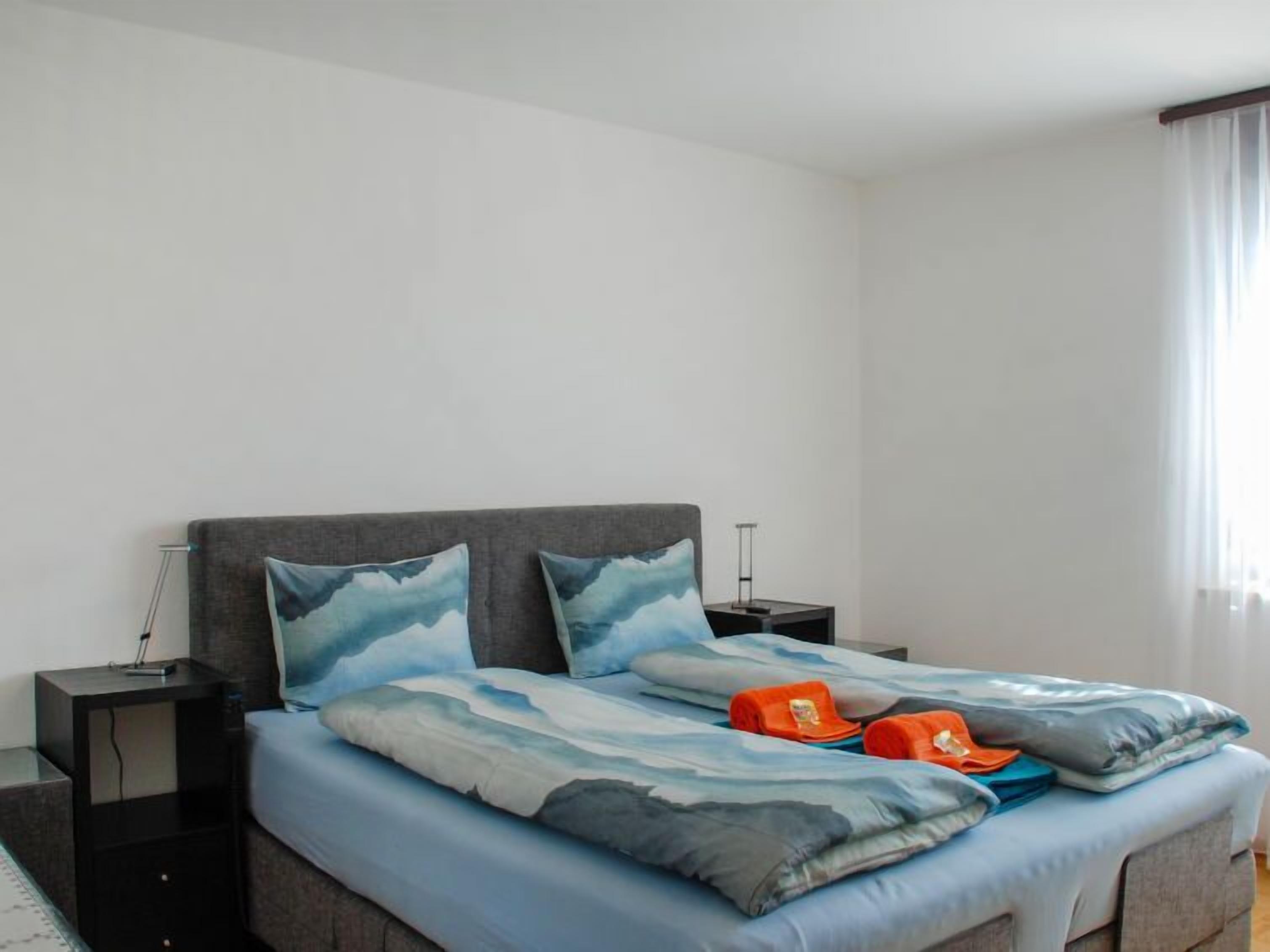 A neatly made bed with a blue and white ocean-themed bedding set, two bedside tables with lamps, and a window with sheer curtains.