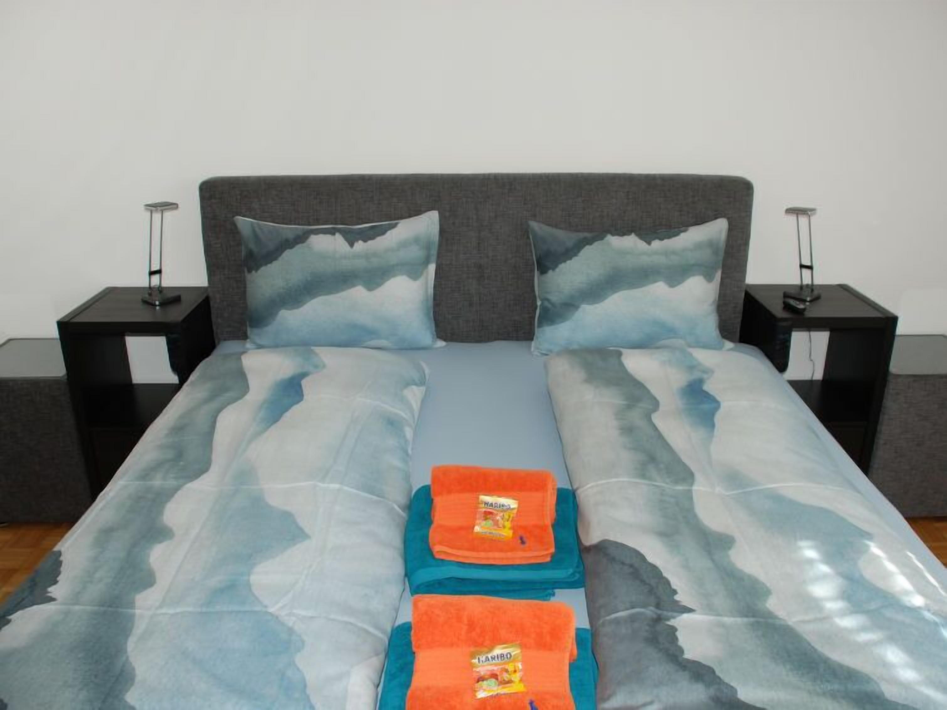 A bed with a grey headboard, two matching pillows, and a blue and white patterned bedspread. There are two black nightstands with lamps on either side of the bed.