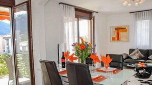 Ein modernes Wohnzimmer mit einem Glastisch, schwarzen Stühlen und einem Sofa. Ein großes Fenster mit Vorhängen, ein Gemälde an der Wand und Blick auf Gebäude im Freien.