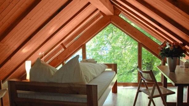 Un dormitorio acogedor en el ático con techo inclinado de madera, una cama con almohadas, una pequeña mesa con un jarrón y una silla.