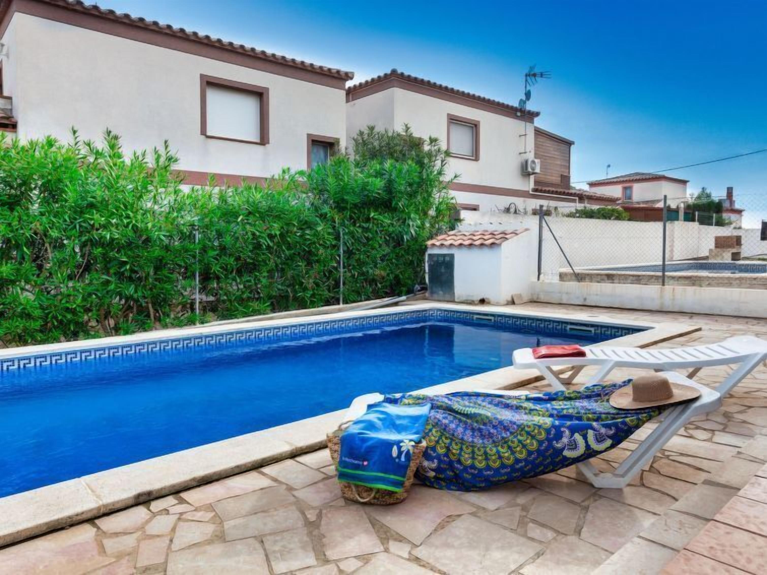 A swimming pool with a tiled deck, a lounge chair, and a covered area with a patterned fabric.