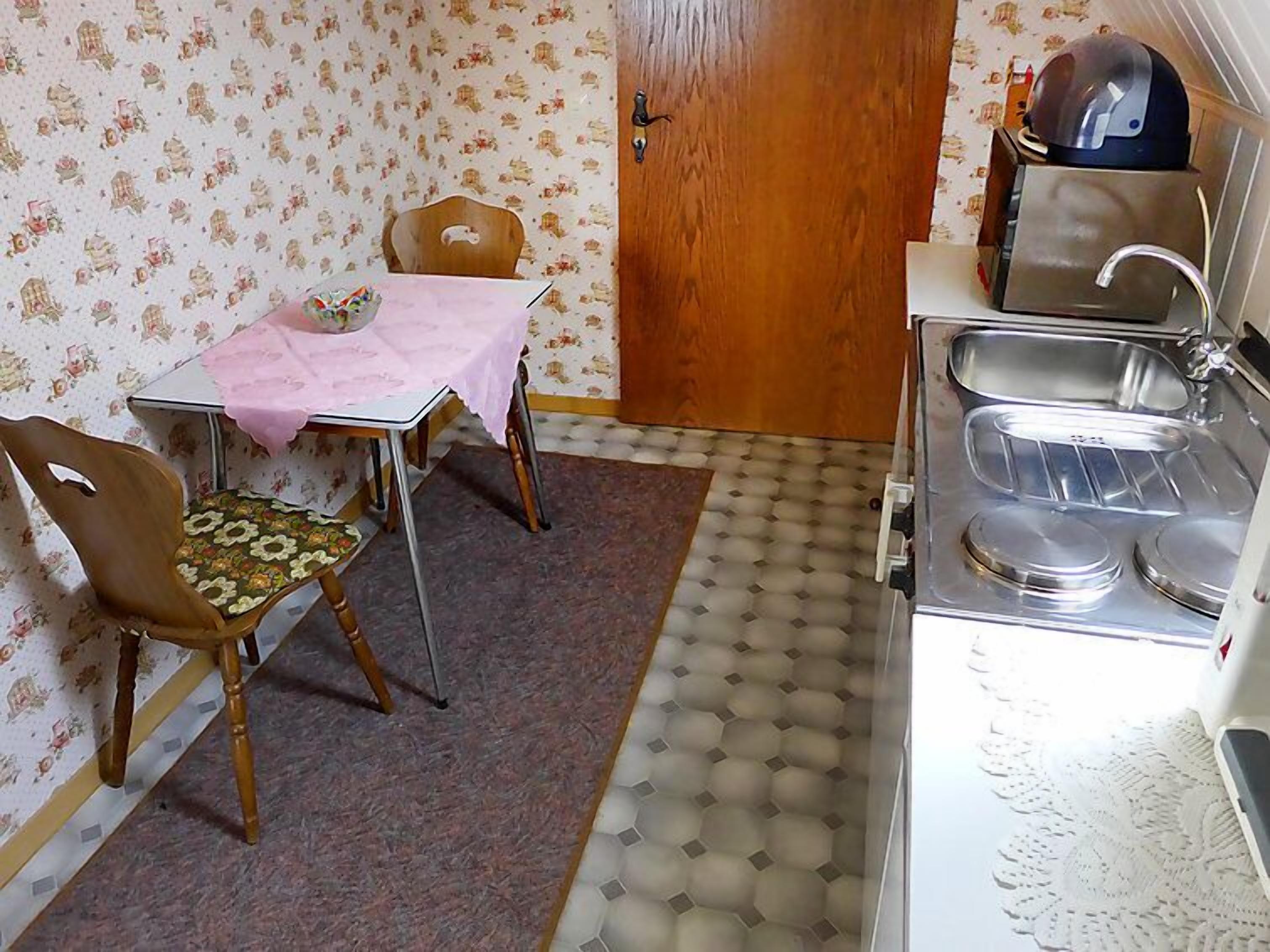 A compact kitchen with a wooden door, a table with a pink tablecloth, two wooden chairs, and a sink with a kettle.
