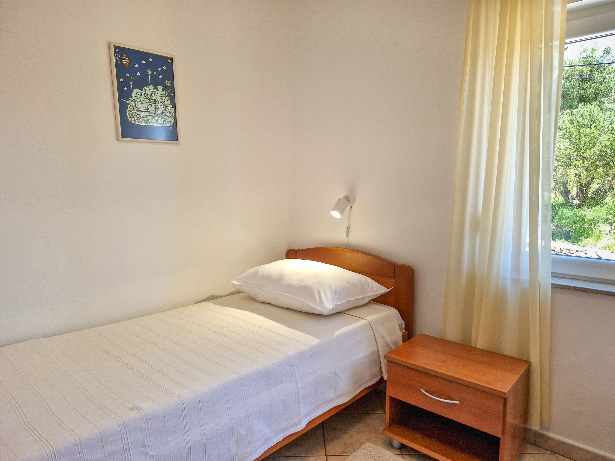 Una habitación pequeña con una cama individual, cabecera de madera, mesita de noche y una lámpara de pared. Hay una ventana con cortinas y un cuadro enmarcado en la pared.