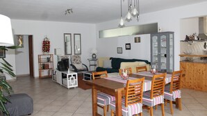 A dining area with a wooden table and chairs, a sofa, a TV, and a cabinet in a well-lit room with white walls.