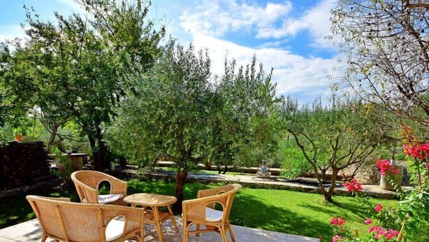 Un'area patio esterna con mobili in vimini e pavimento in pietra, circondata da vegetazione lussureggiante e piante fiorite.