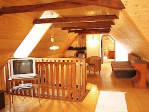 A cozy attic room with a wooden ceiling, a television on a wooden stand, a cushioned chair, and a small table.