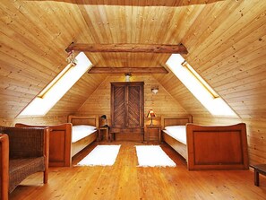 A cozy attic bedroom with wooden walls and a sloped ceiling. There are two single beds with white linens, a wicker chair, and a small table with a lamp.
