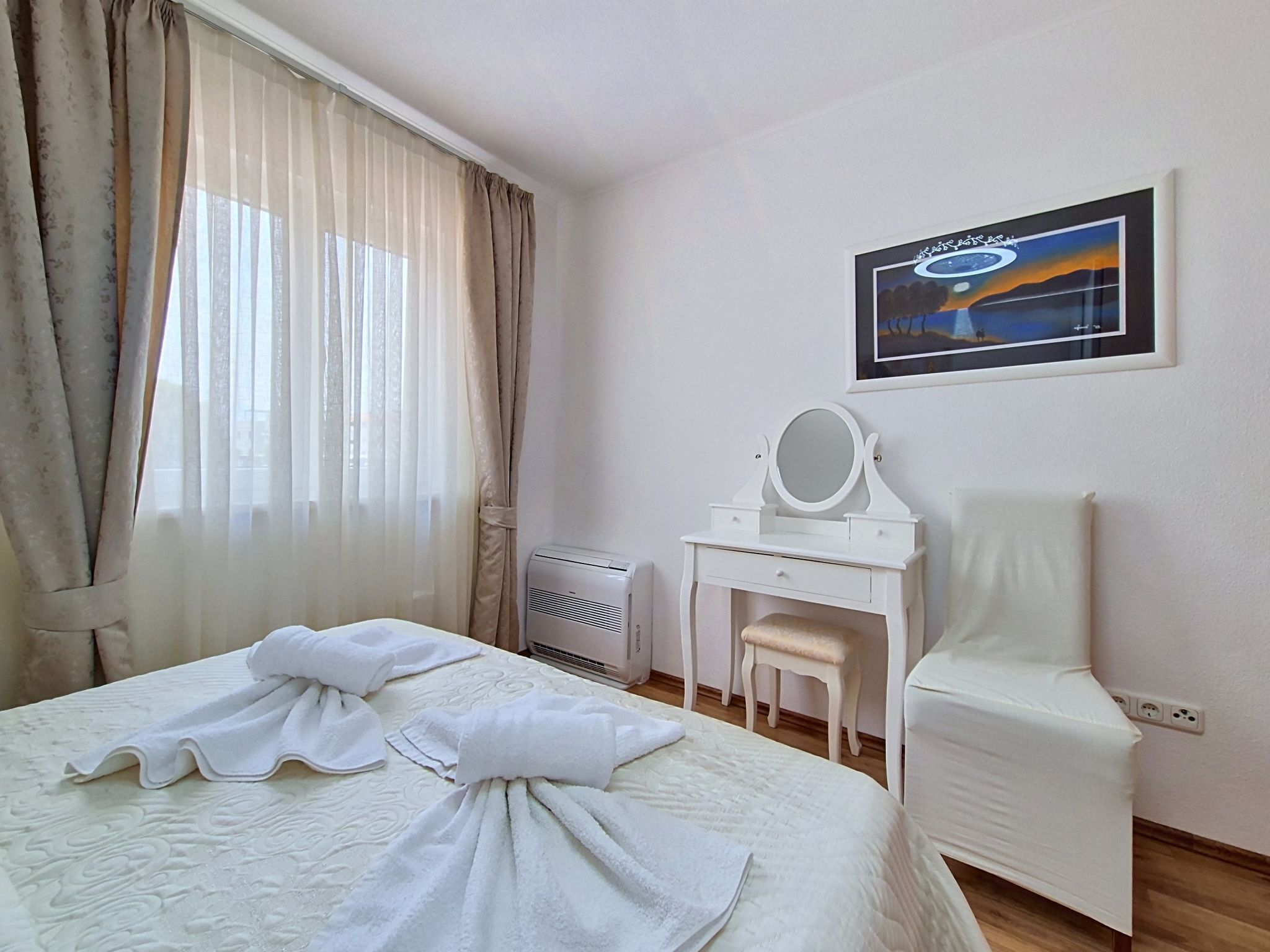 Quarto de hotel com uma cama, uma penteadeira branca com espelho, uma cadeira e um quadro na parede.