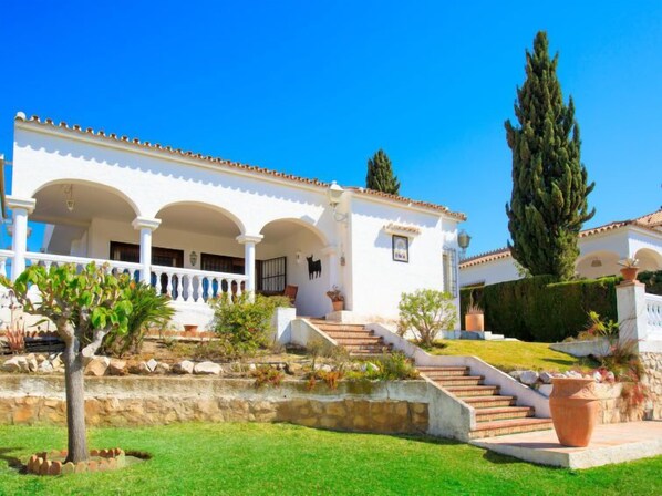A white building with a covered porch, stone steps, and a small garden with a palm tree.