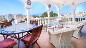Eine Terrasse mit einem runden Tisch und Korbstühlen, mit Blick auf eine Stadtlandschaft und das Meer.