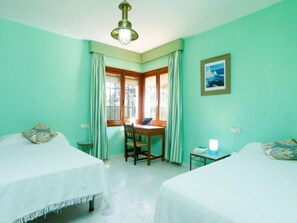 A room with two beds, a desk, and a chair. There is a window with curtains, a picture on the wall, and a ceiling light fixture.