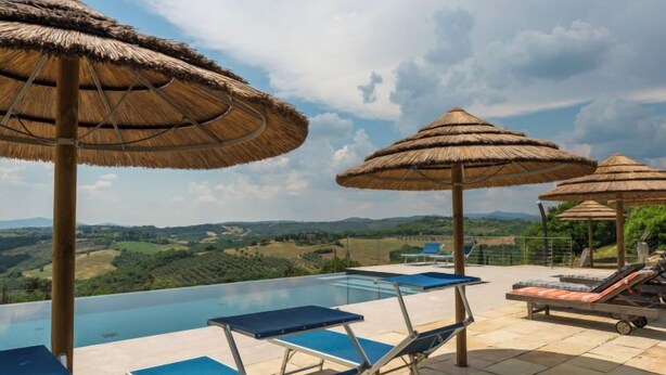 Une piscine à débordement avec des chaises longues bleues et des parasols en chaume, offrant une vue imprenable sur un paysage magnifique.