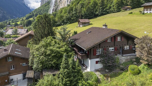 A chalet-style building with a sloped roof, surrounded by lush greenery and a mountain backdrop.