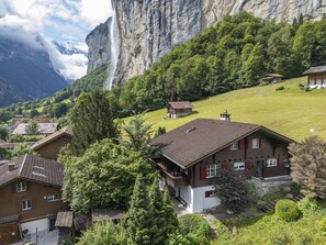 Un edificio in stile chalet con tetto spiovente, circondato da vegetazione rigogliosa e con le montagne sullo sfondo.