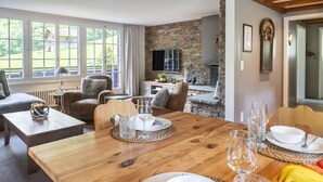 A dining area with a wooden table set for four, a stone fireplace, and a view of the outdoors through large windows.