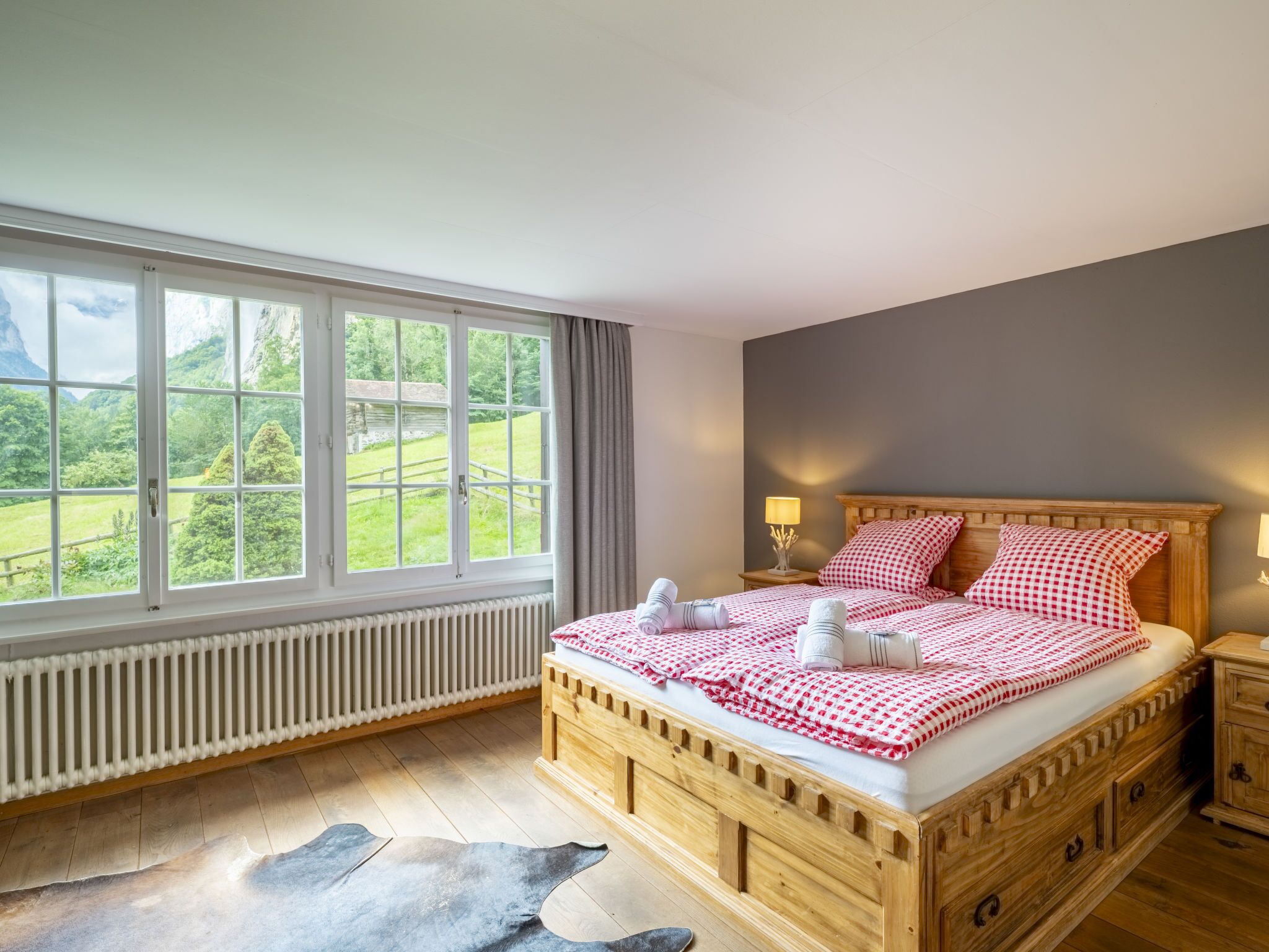 Ein hölzernes Bett mit karierter Bettdecke, ein Nachttisch mit Lampe und Blick auf eine grüne Landschaft durch große Fenster.