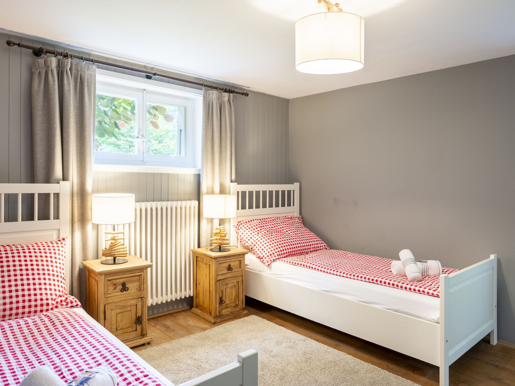 A bedroom with two single beds, a wooden nightstand with a lamp, a wooden dresser, and a window with curtains.