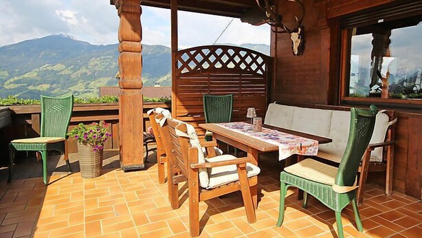 An outdoor patio with wooden furniture, a table with a floral tablecloth, and a view of mountains.