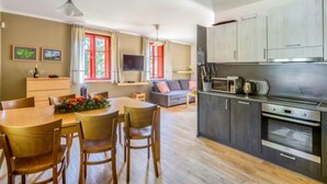 A modern kitchen with stainless steel appliances, a dining area with wooden chairs and a table, and a living area with a sofa and TV.