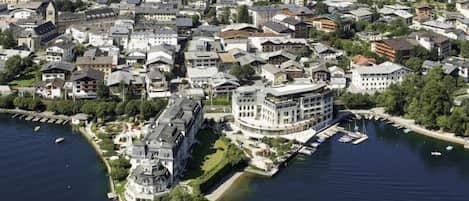 An aerial view of a coastal town with a prominent waterfront property featuring a large, multi-story building with a curved facade, a dock, and a marina with several boats.