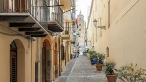 A narrow street with buildings on either side, featuring balconies, potted plants, and a cobblestone path. - Jancaliva by Interhome (Cefalù)