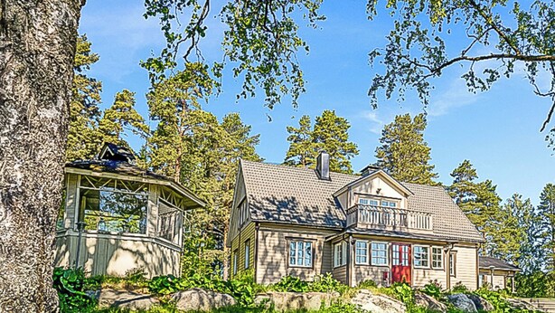 Ett tvåvåningshus med en röd dörr, omgivet av en välskött trädgård och stora stenar.