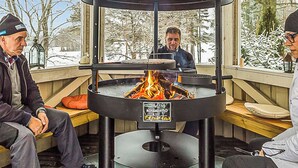 Ett takförsett utomhusområde med sittplatser, en central eldstad, bänkar och utsikt över snötäckta träd.