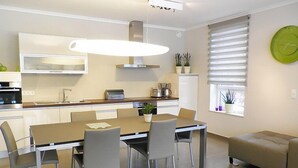 Une cuisine moderne avec un coin repas, comprenant une table avec des chaises, un four encastré et une grande fenêtre avec des stores.