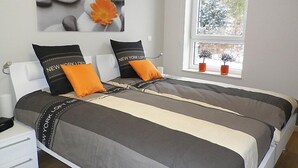 Un lit double avec une literie aux tons gris et beige, deux oreillers ornés de la mention « New York Loft », une fenêtre avec des plantes et un tableau représentant des pierres et une fleur.