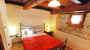 Quarto com cama, mesa de cabeceira, luminária, parede de pedra e janela.