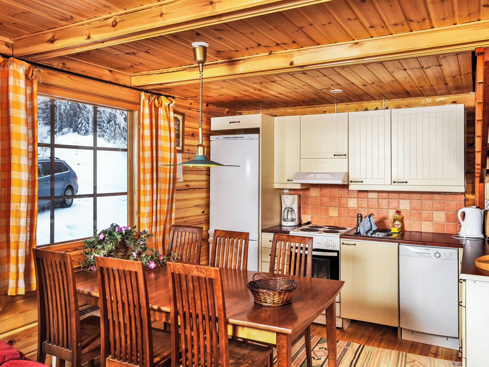 Interior acogedor de una cabaña de madera, con comedor, cocina y vistas a un paisaje nevado a través de la ventana.
